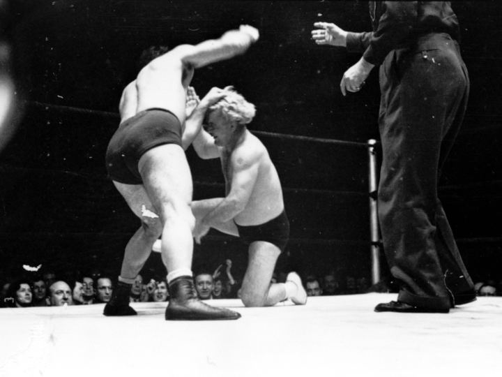 Black and white 1949 photo of Gorgeous George kneeling during a wrestling match